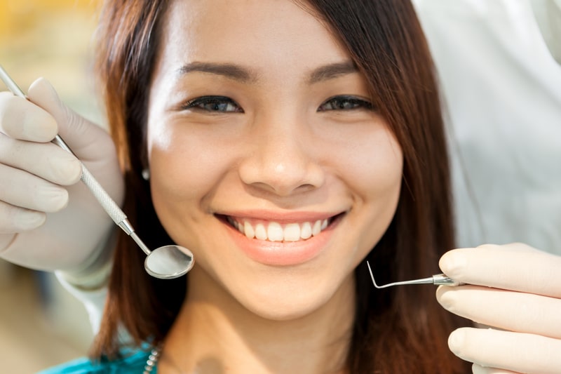 A beautiful brunette young adult that is smiling as a dental assistant is about to examine her mouth with dental tools. 