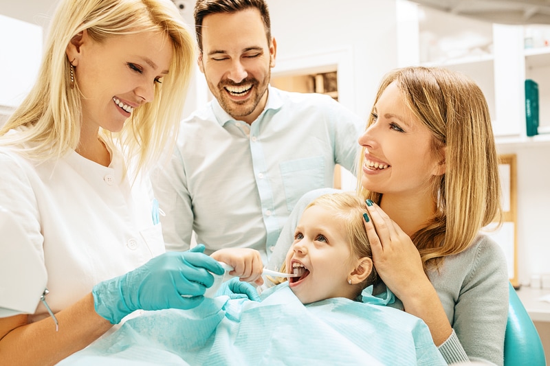 A dental hygienist helping a small child to brush their teeth while the mother and father are right there with the child smiling.