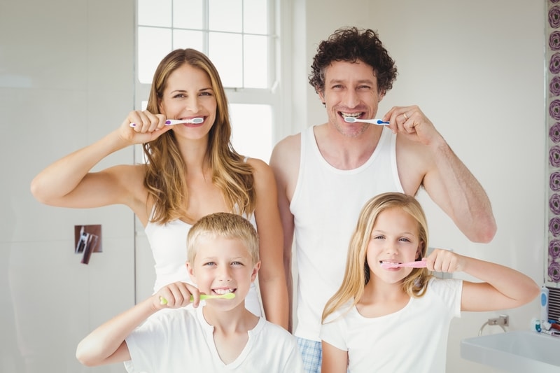 A family of four with one man and one woman and one young boy and young girl. All are wearing white to suggest cleanliness and they are brushing their teeth.