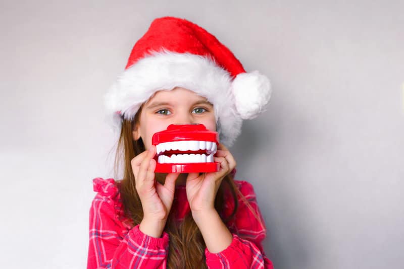 Little girl wearing a santa hat and holding fake teeth in front of her mouth.
