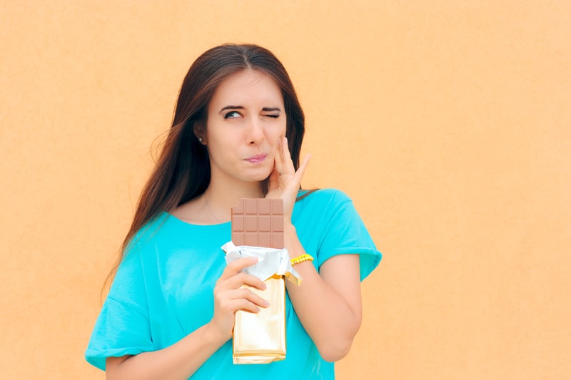 Girl that is holding her face and eating a chocolate bar, but she is having tooth pain. 