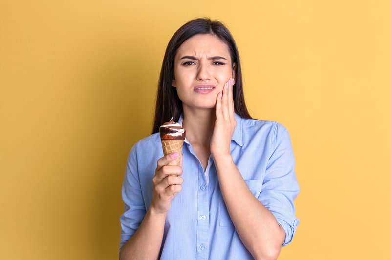 Woman standing with an ice cream cone in her hand while holding her jaw in pain.